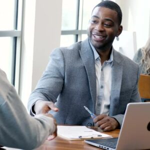Mid adult male bank employee uses a laptop before shaking hands in greeting with a senior male customer. They then begin to discuss the bank's services